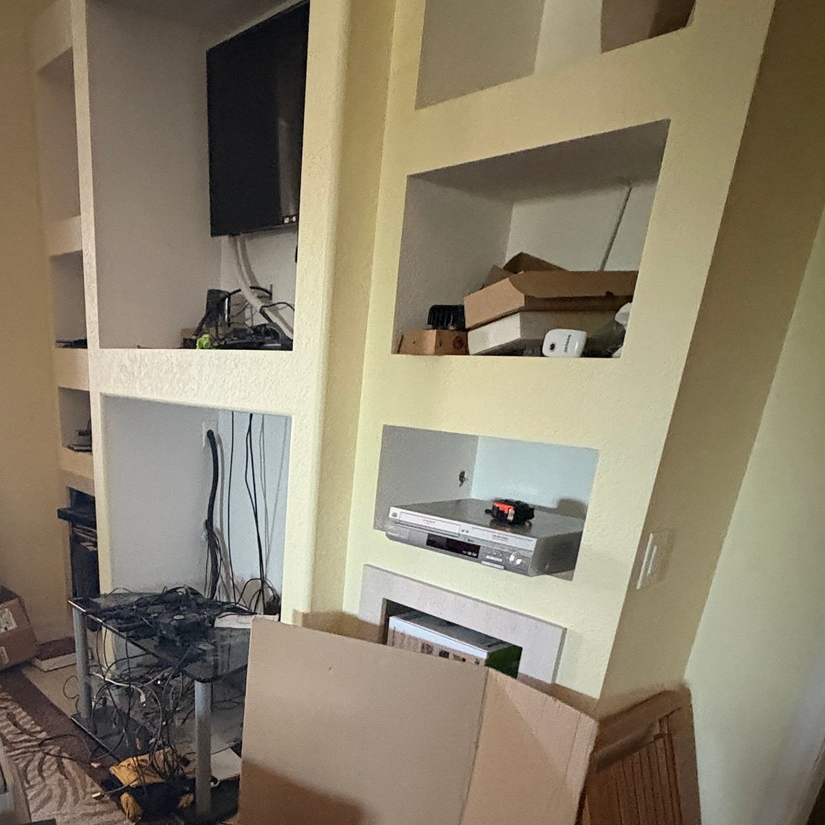 Before: a living-room built-in wall crowded with a TV, boxes and electronics on the shelves, a glass TV stand, flattened cardboard and a patterned rug in front.